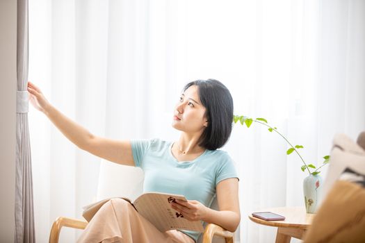 A woman relaxes in a bright living room, holding a book near a coffee table with a vase.