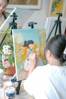 A woman painting a vibrant artwork on canvas indoors with art supplies and a flower pot nearby.
