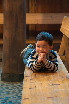 A cheerful young boy lying on a wooden bench in a cozy indoor setting.