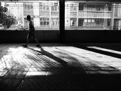 A person walks through a shadow-filled corridor in a Hong Kong urban setting, captured in black and white.
