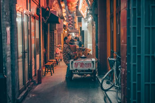 A bustling night scene in a lively Beijing alleyway with street vendors and colorful lanterns illuminating the path.