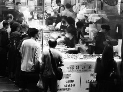 Black and white image of a busy street food stall in Hong Kong with people queuing.