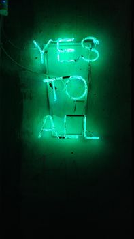 Bright green neon sign displaying 'Yes to All' against a dark wall background.