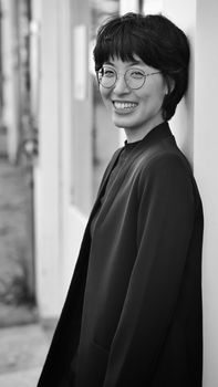 Black and white portrait of a young woman in a blazer and eyeglasses smiling outdoors.