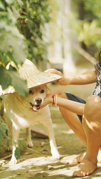 A playful scene of a dog wearing a straw hat with a woman's hand on its face, outdoors.