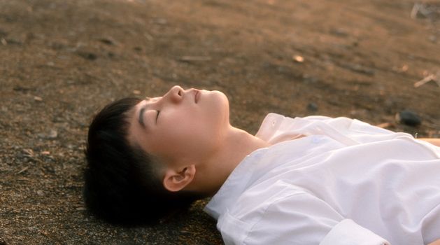 A young man in a white shirt laying peacefully on the ground outdoors.