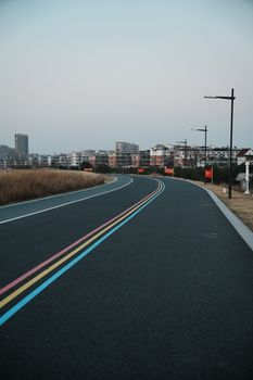 A serene urban road with colored lines, bordered by fields and city buildings at sunset.