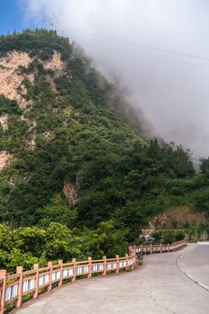 A winding road leads through lush greenery towards a mist-covered mountain, evoking a serene and quiet atmosphere.