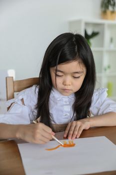 From above of concentrated little Asian schoolgirl in white shirt drawing on paper sheet with watercolor using paintbrus