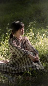 Calm moment of a young Asian woman in plaid dress sitting in lush grass, embraced by sunlight.